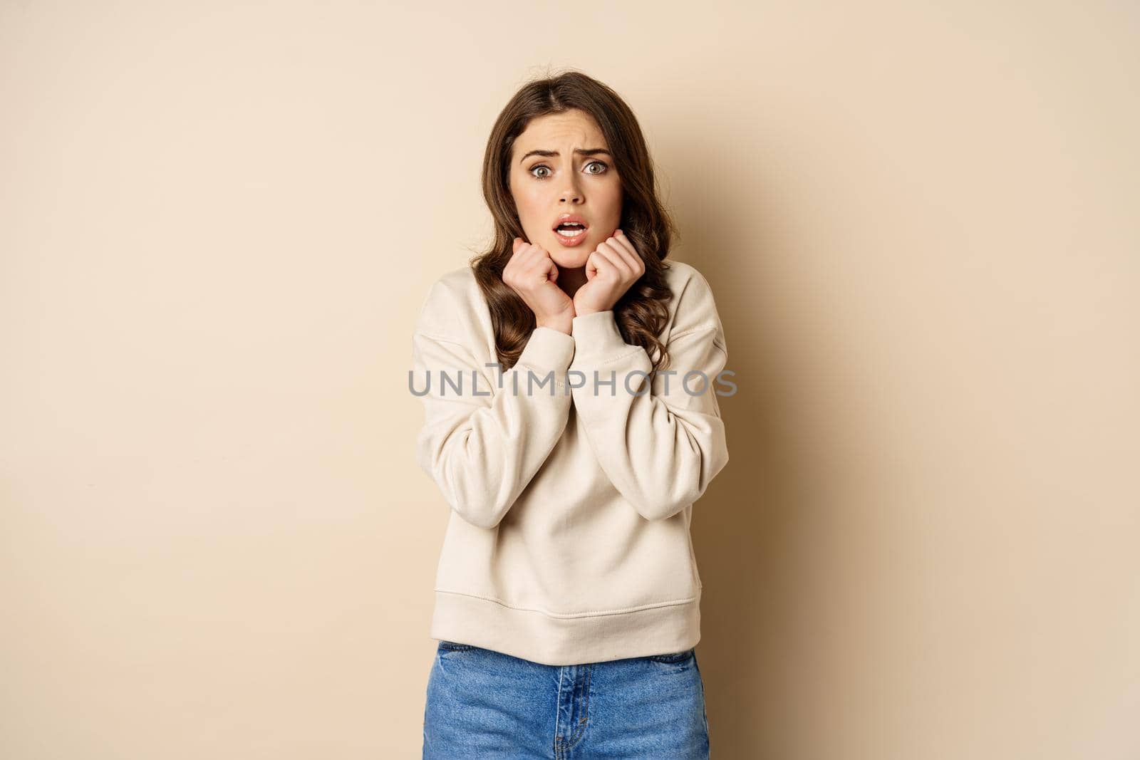 Scared insecure woman trembling from fear, looking at camera shocked and frightened, standing over beige background.