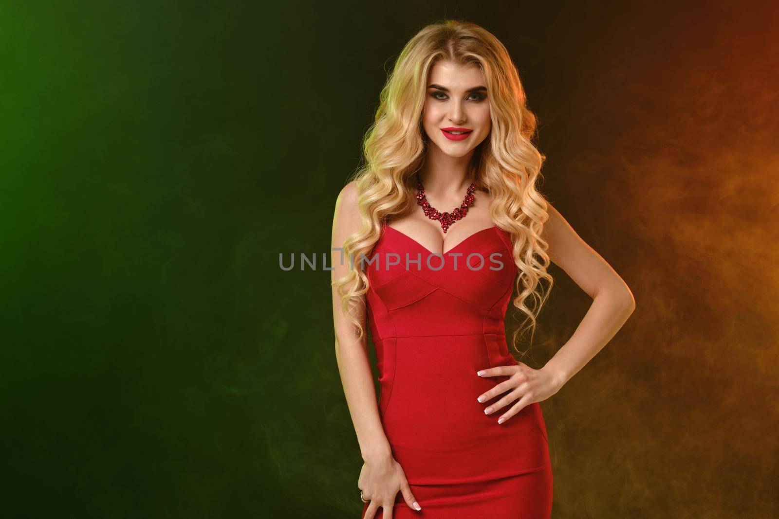 Curly blonde woman with bright make-up and alluring decollete, in red fitting dress and necklace. Smiling, hand on waist, posing on colorful smoky background. Fashion and beauty. Close up, copy space
