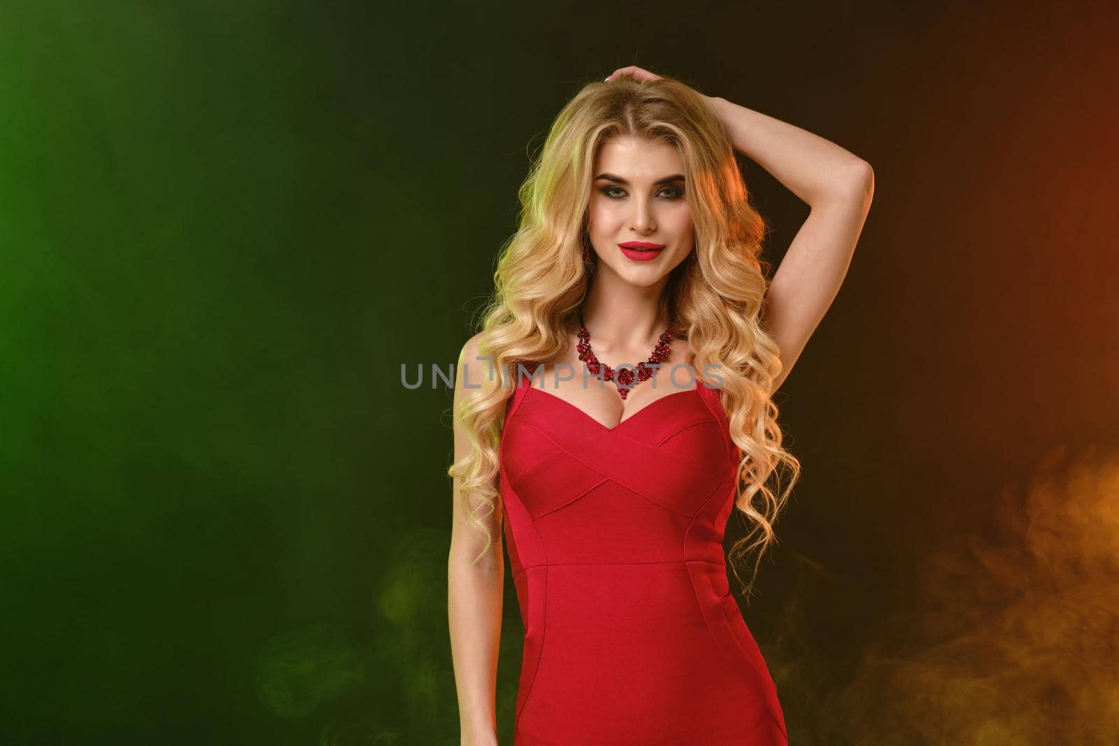Charming curly blonde lady with bright make-up, in red fitting dress and necklace. She is smiling while posing against colorful smoky studio background. Fashion and beauty. Close up, copy space