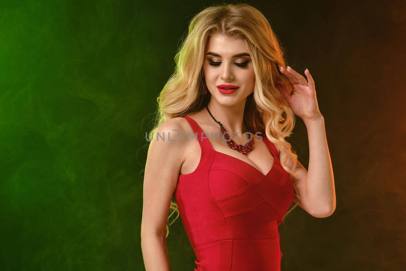 Charming curly blonde female with bright make-up, in red fitting dress and necklace. She is smiling, looking down, posing on colorful smoky studio background. Fashion and beauty. Close up, copy space