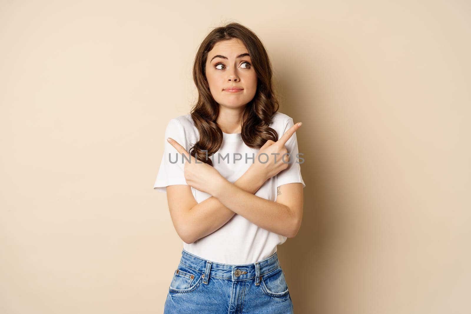 Beautiful girl choosing between two options, pointing sideways and looking clueless, standing over beige background.