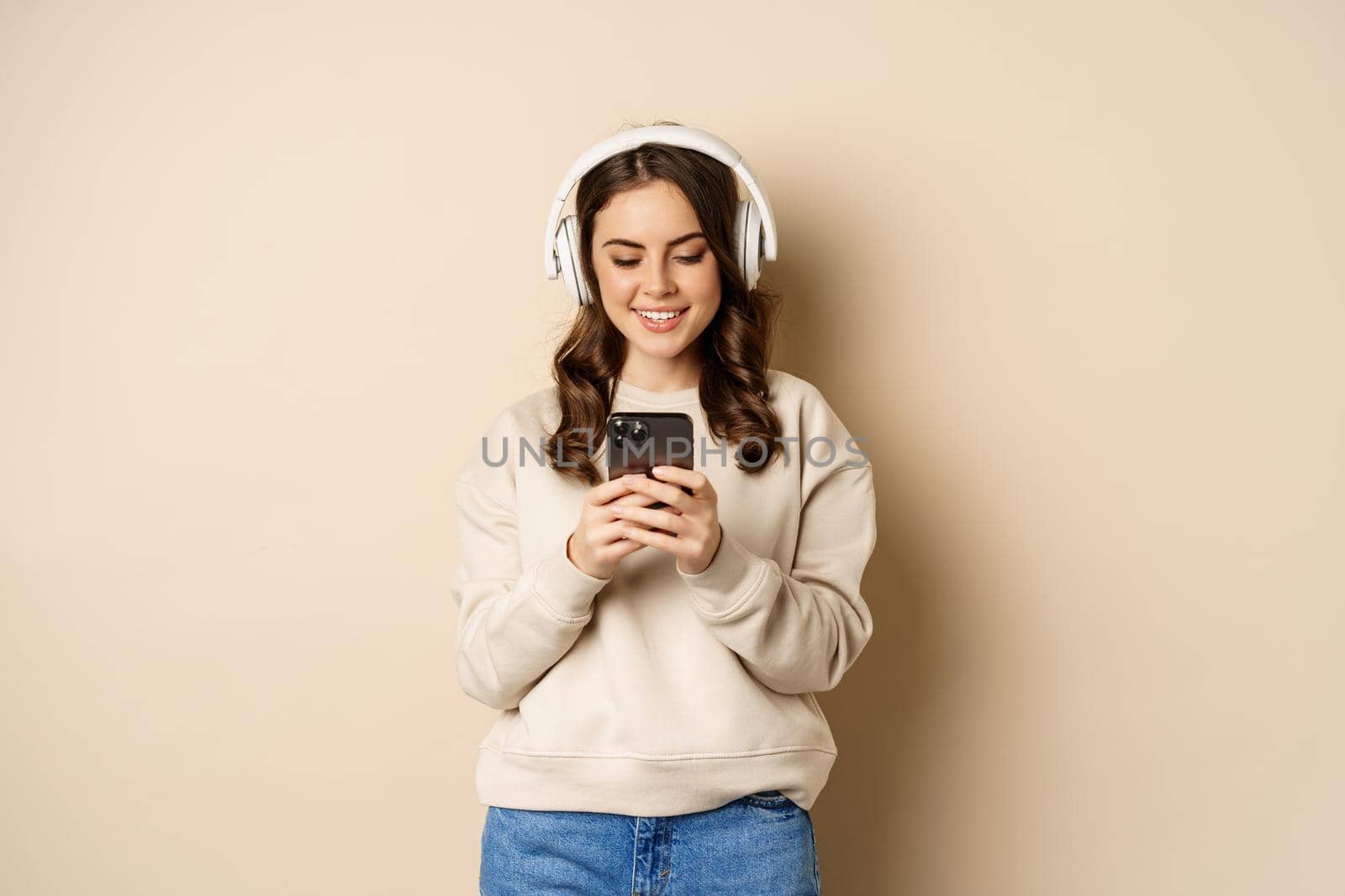 Smiling cute girl in headphones, looking at mobile phone, listening music or podcast, standing over beige background.