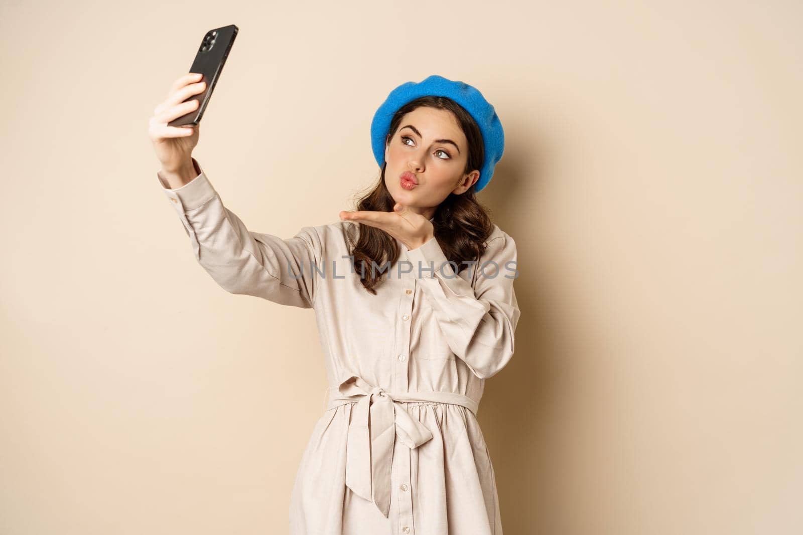 Beautiful modern woman posing for selfie, holding smartphone, taking picture photo on mobile phone, standing happy against beige background by Benzoix