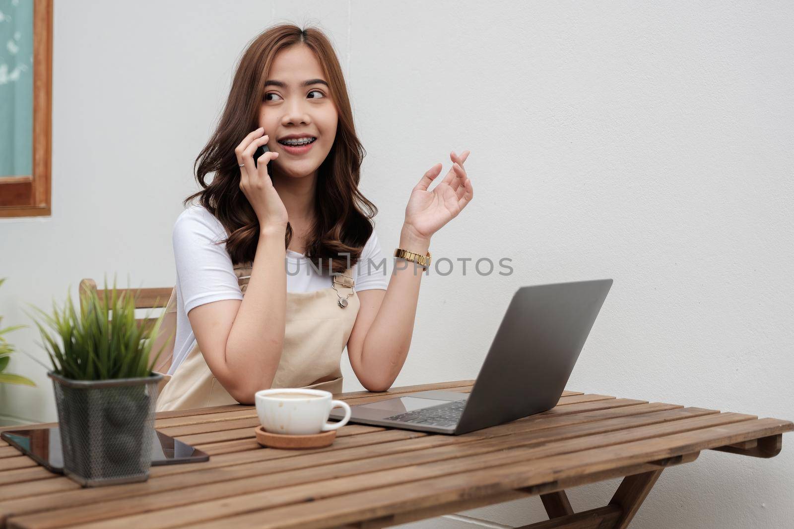 Smiling young woman talking on the phone while using laptop at cafe shop, happy young girl holds cellphone making answering call, attractive teenager having pleasant conversation chatting by mobile with friend.