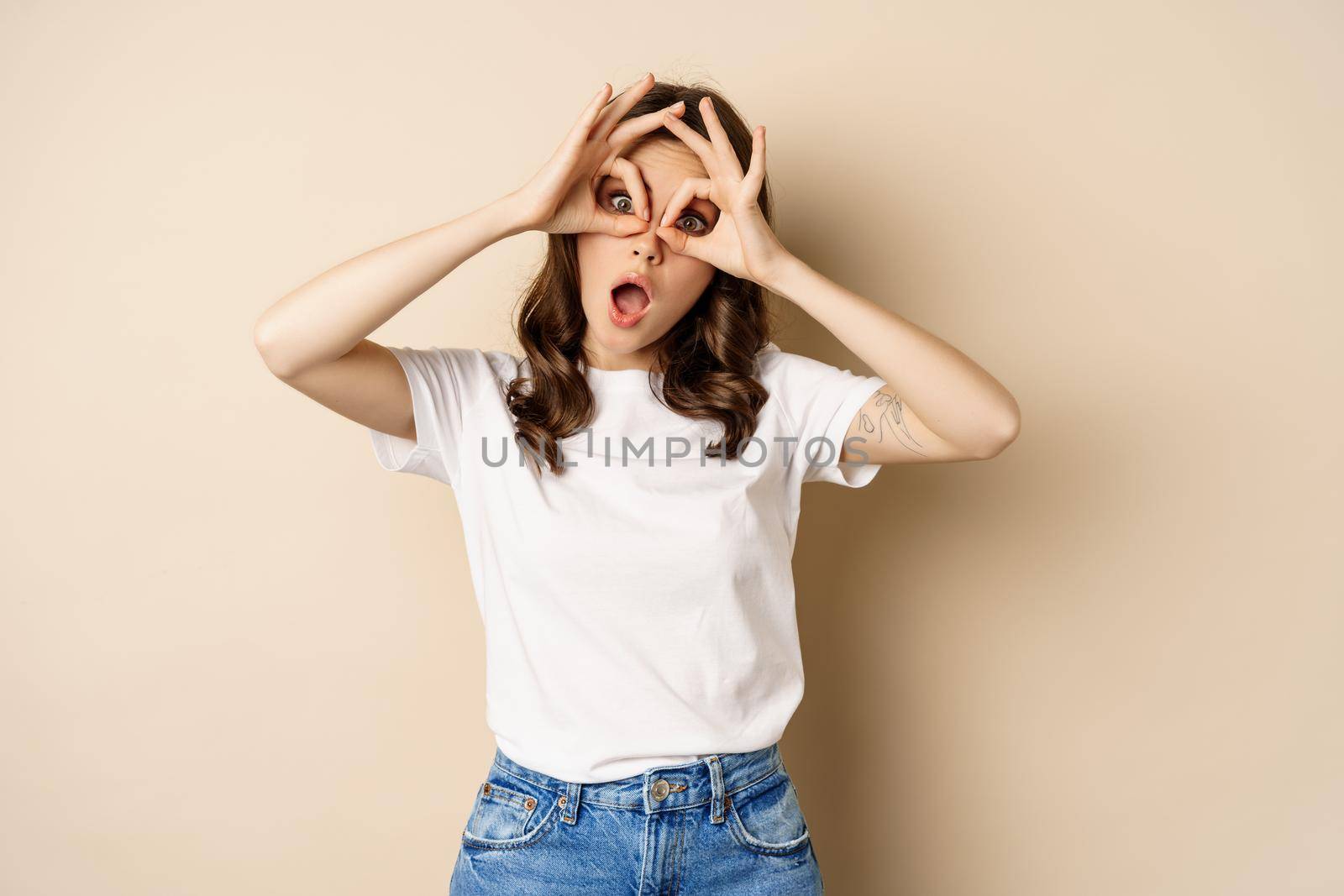 Funny brunette woman having fun, showing finger glasses gesture and fool around, posing over beige background.