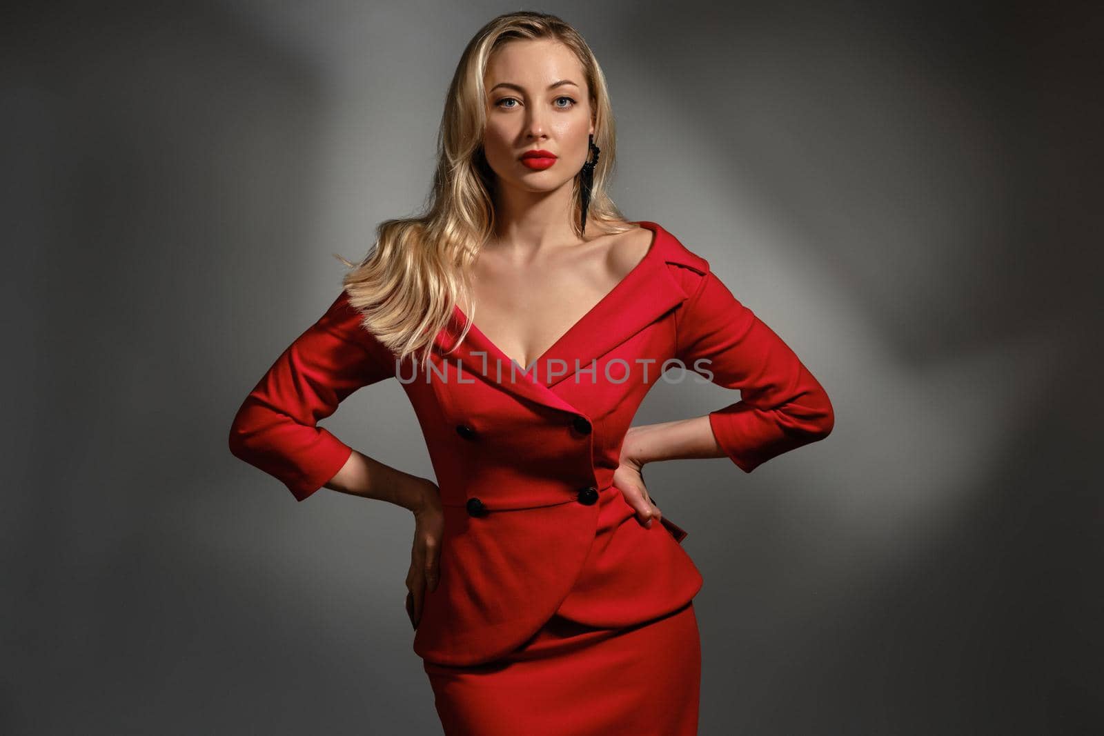 Gorgeous blonde lady, bright make-up and bare shoulders, in red stylish dress and black earrings. Hands on waist, posing against gray studio background. Fashion and beauty. Close up, copy space