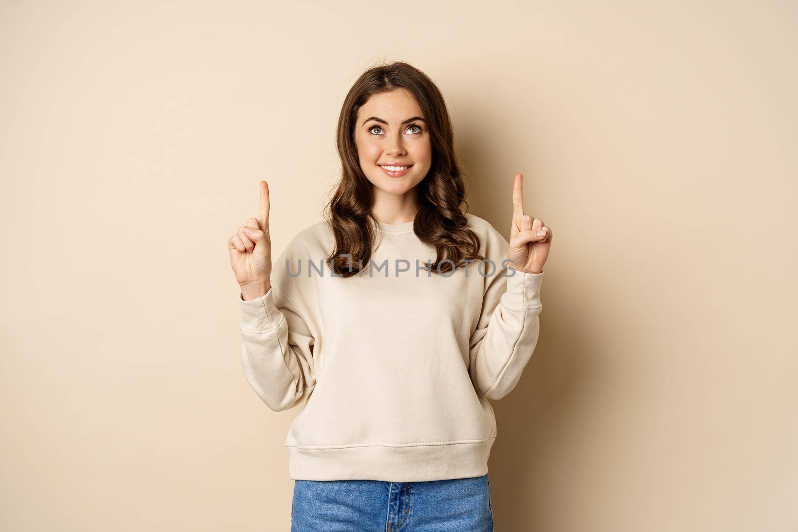 Enthusiastic girl pointing fingers up, showing announcement, promo text or logo upwards, standing over beige background.