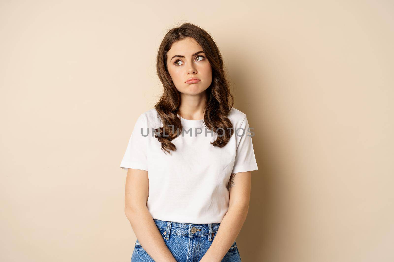 Sad and tired brunette woman looking drained and unamused, standing with pokerface against beige background.