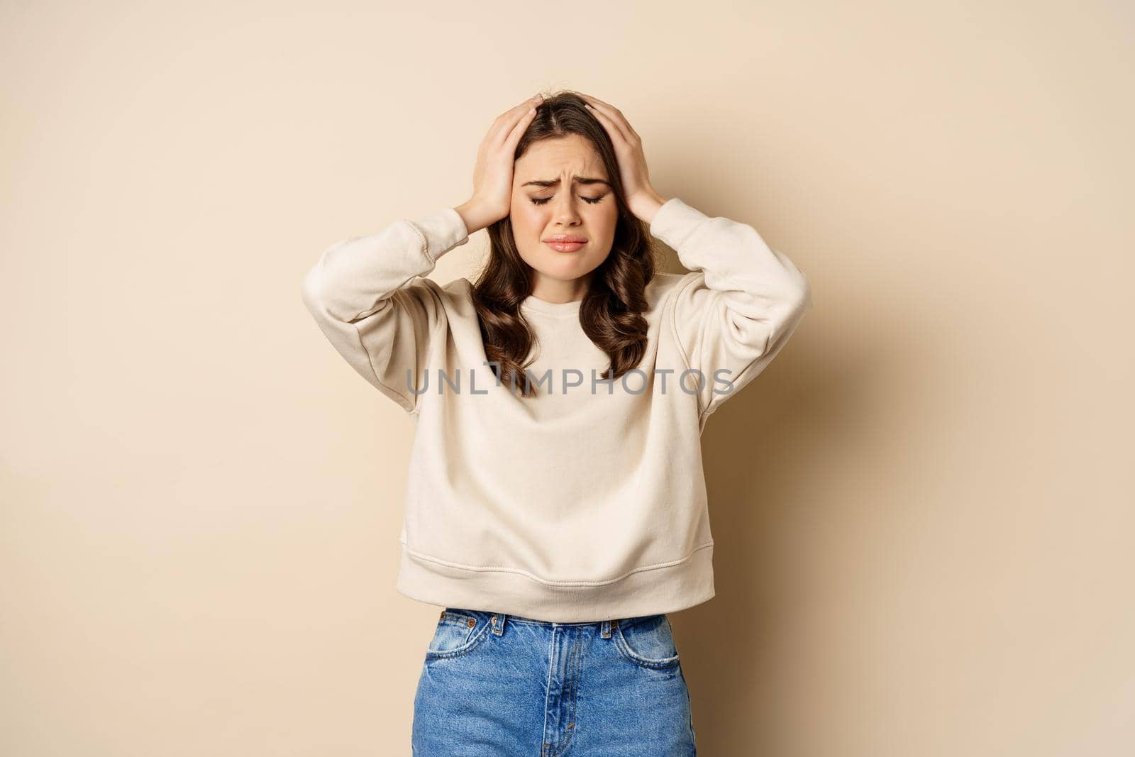 Frustrated and troubled woman facing disaster, holding hands on head in panic, looking anxious, standing over beige background.