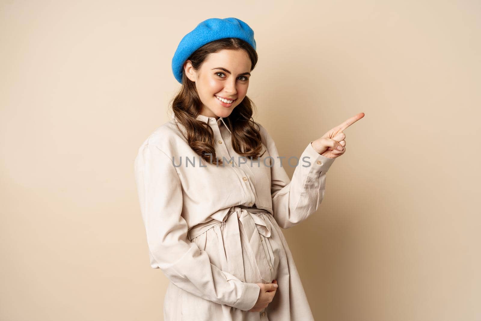 Young future mother, pregnant woman smiling, posing with big belly third trimester of pregnancy, looking happy, expecting baby, pointing right at logo, banner or advertisement.