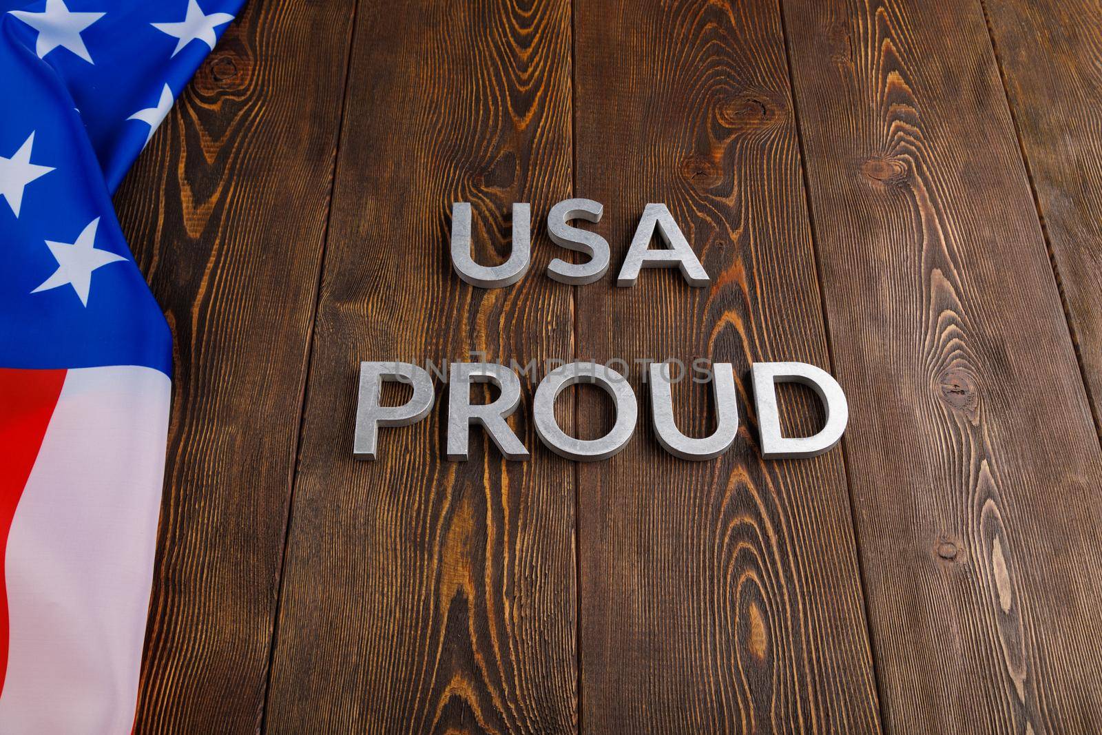 words USA proud laid with silver metal letters on brown wooden surface with flag of United States of America.