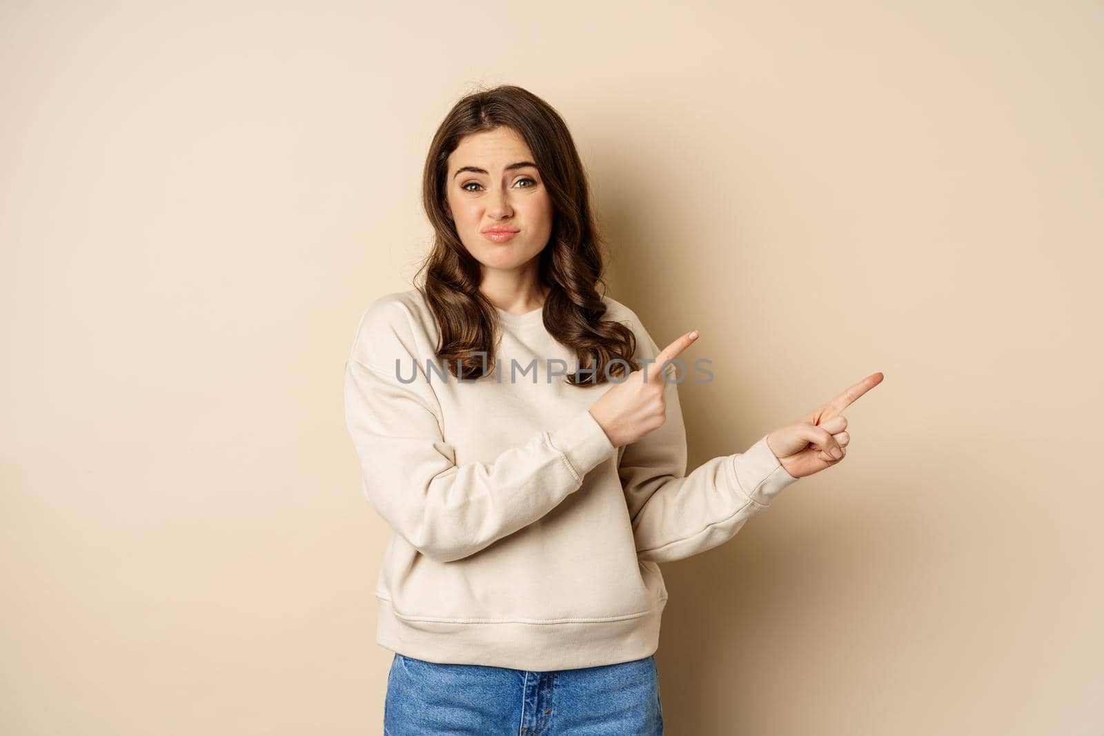 Skeptical girl looking hesitant, pointing fingers right at logo, promo text, standing over beige background.
