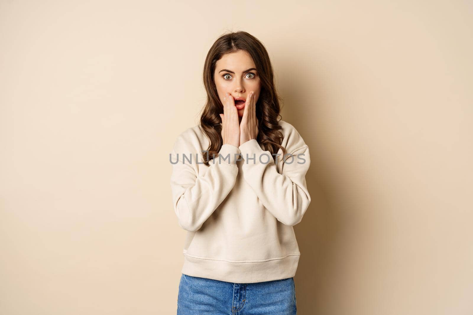 Portrait of shocked brunette woman drop jaw, gasping and staring speechless at camera, beige background. Copy space