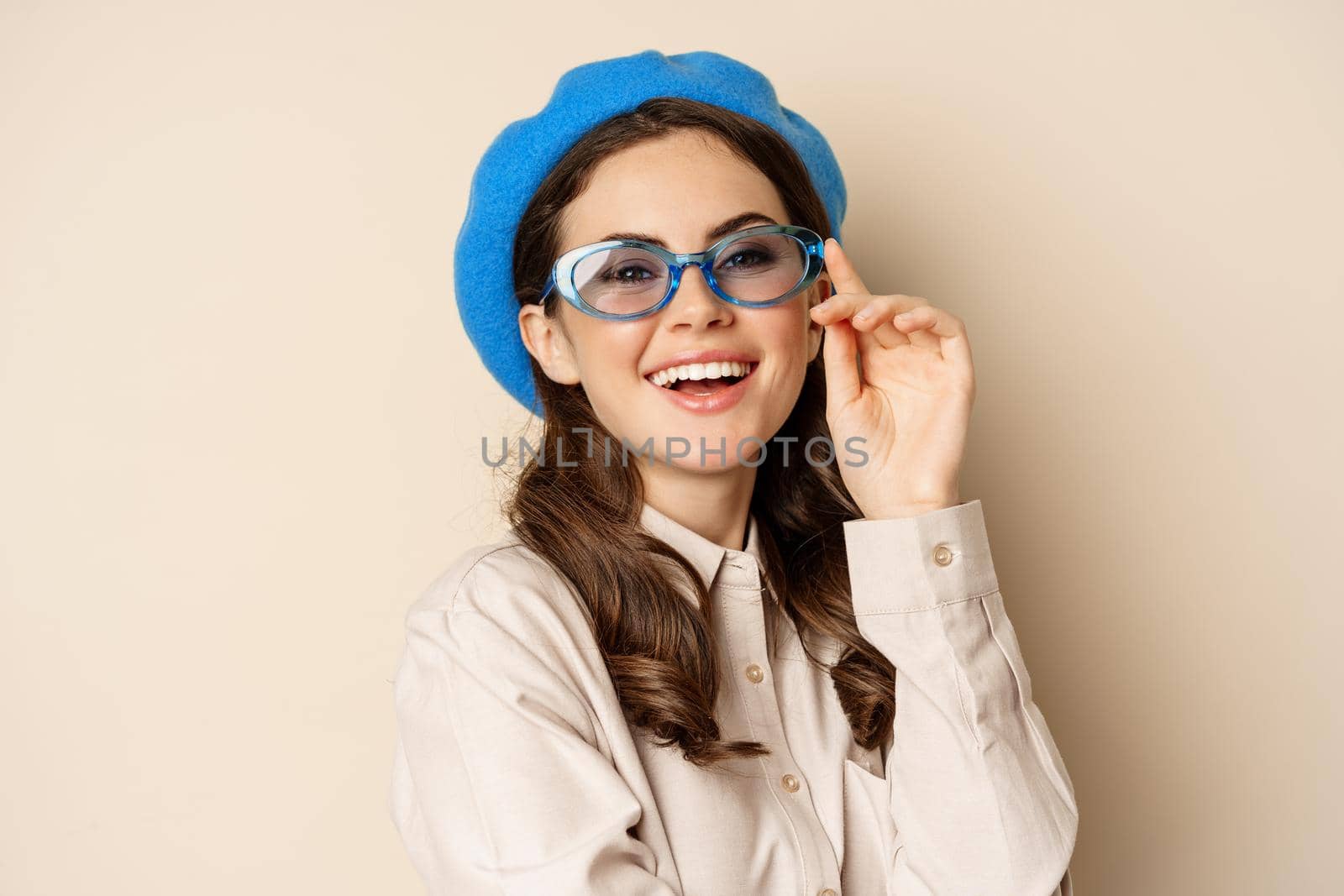 Close up portrait of stylish woman posing in sunglasses, laughing and smiling, wearing trendy outfit, standing over beige background.