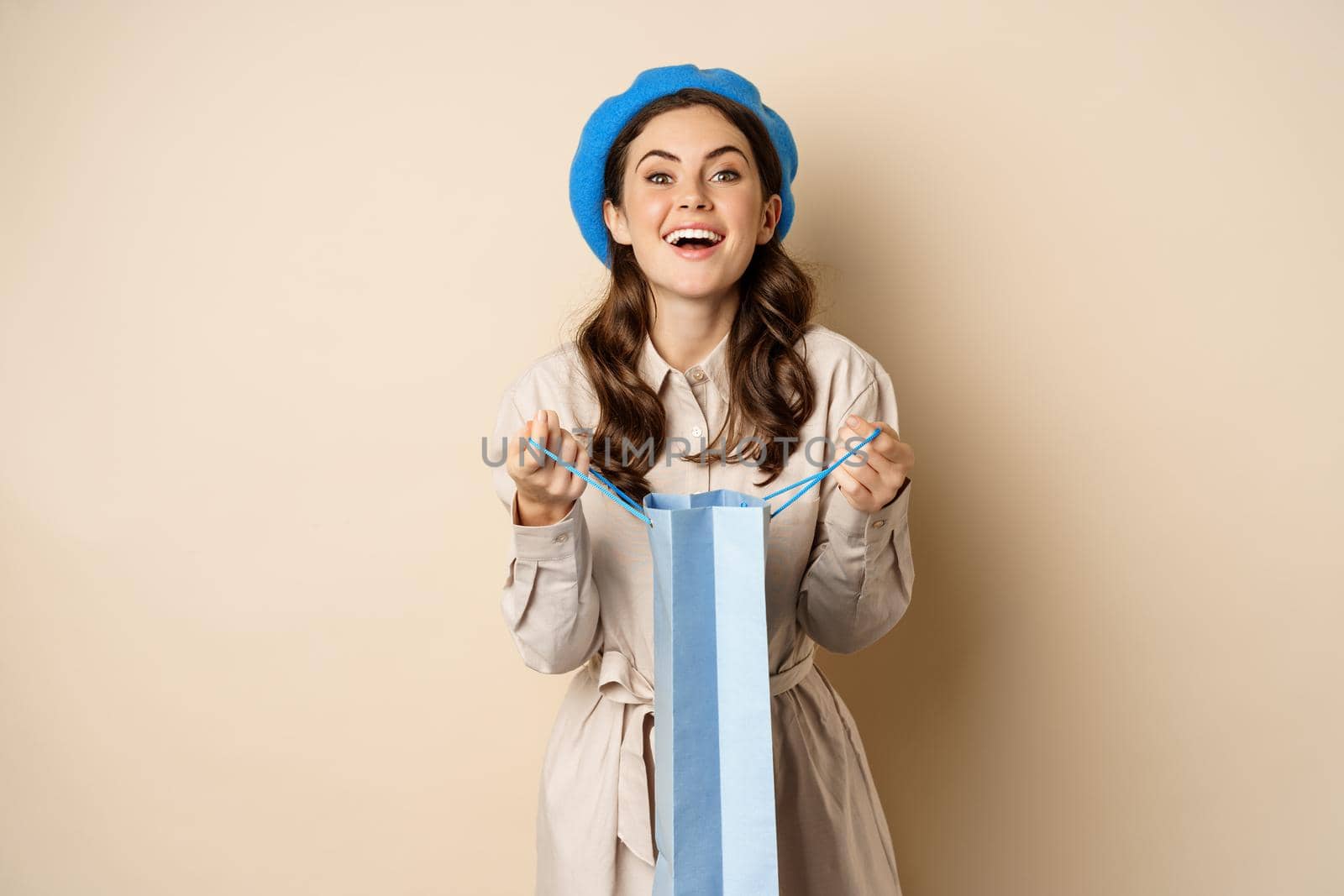 Happy young woman smiling, open shopping gift bag and laughing, giving present, standing over beige background.