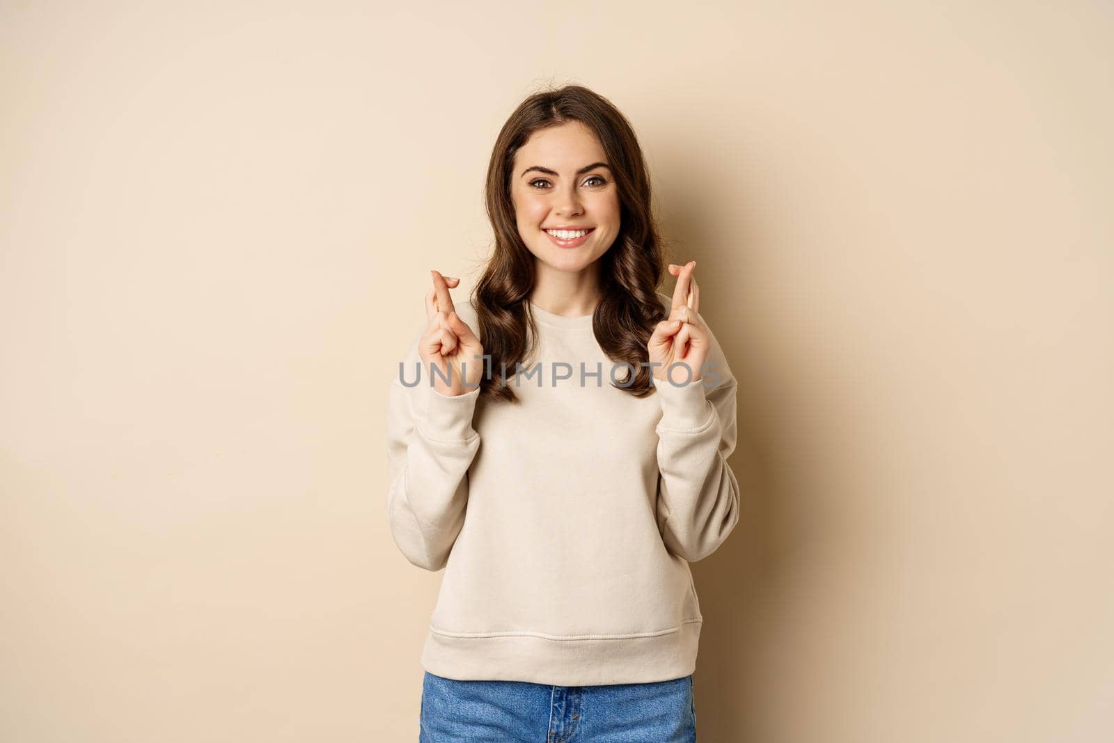 Confident woman praying, making wish with fingers crossed for good luck, standing over beige background.