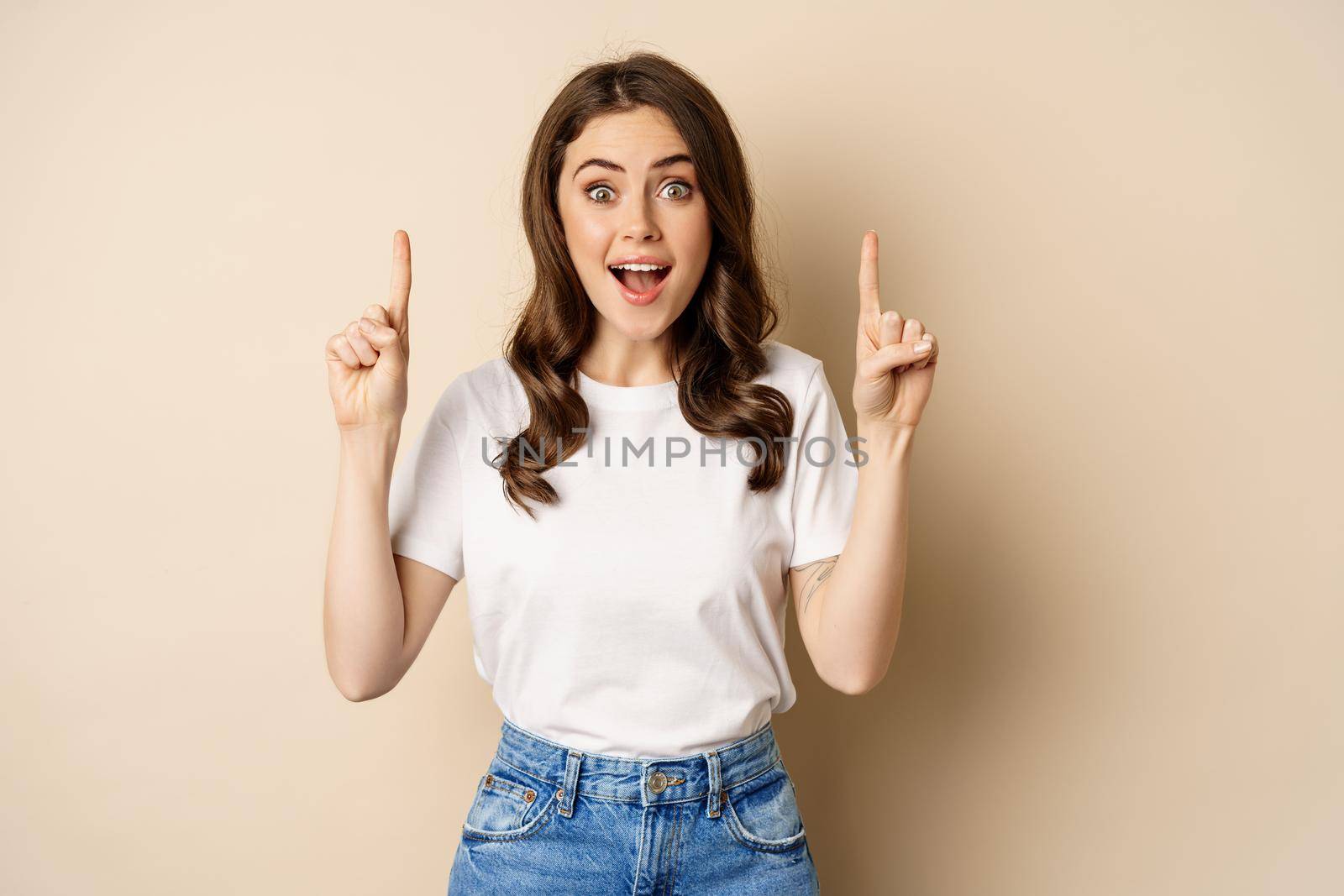 Enthusiastic young woman, female customer pointing fingers up and smiling, showing banner or logo, standing against beige background.