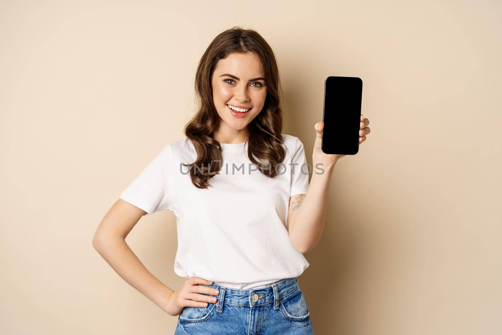 Online shopping and app concept. Young woman smiling and showing mobile phone screen, application interface, standing over beige background.