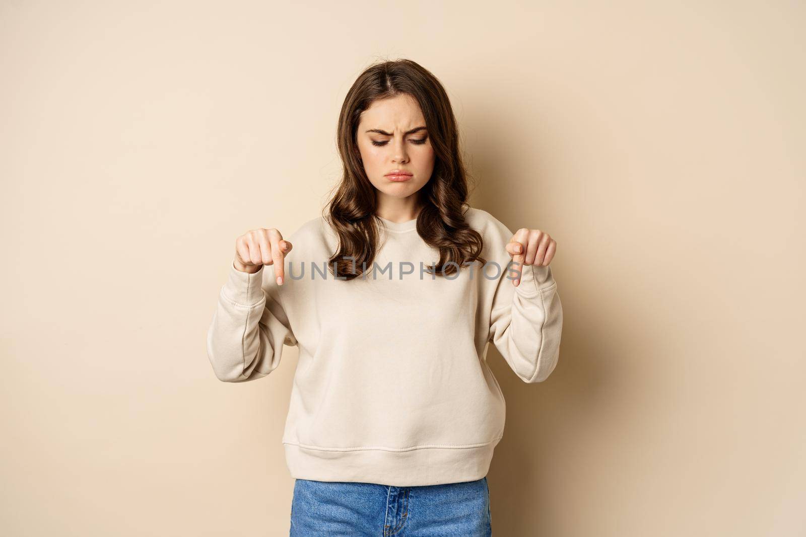 Sad and disappointed girl pointing, looking down with upset grimace moody face, standing over beige background.