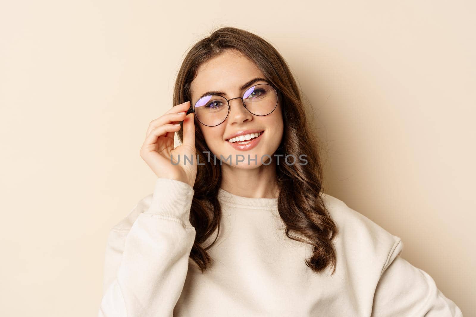 Close up portrait of beautiful modern woman in glasses, smiling and looking happy, posing in eyewear against beige background.