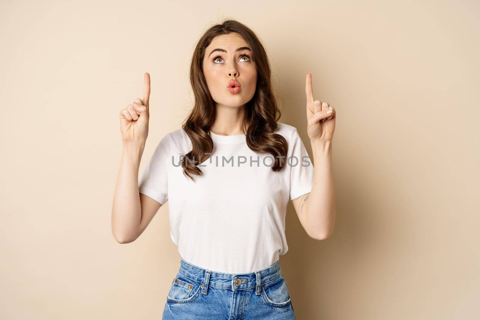 Surprised brunette girl showing promo, pointing and looking up with amazed face expression, standing over beige background.