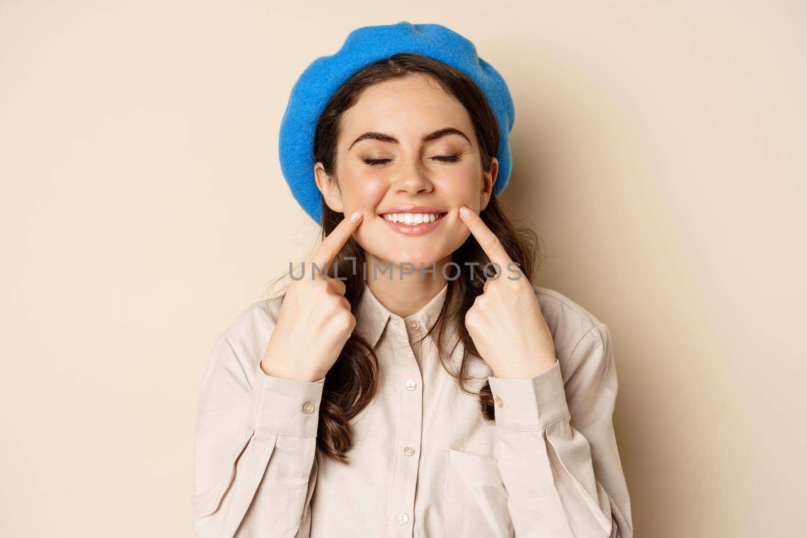 Cute happy woman smiling white teeth, pointing at her dimples, standing over beige background. People and faces concept