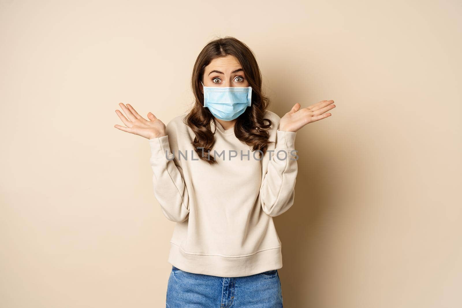 Covid-19, pandemic and quarantine concept. Surprised brunette woman in face medical mask, looking amazed at camera, watching big news, beige background.