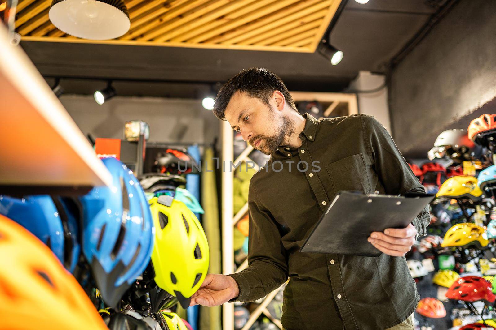 A male bike shop manager makes an inventory of sports helmets in a bike shop. The owner of a sports store with a clipboard in his hands checks the prices of bicycle helmets in the showcase by Tomashevska