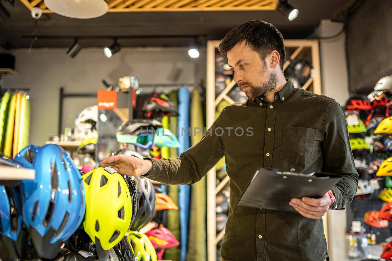 A male bike shop manager makes an inventory of sports helmets in a bike shop. The owner of a sports store with a clipboard in his hands checks the prices of bicycle helmets in the showcase.