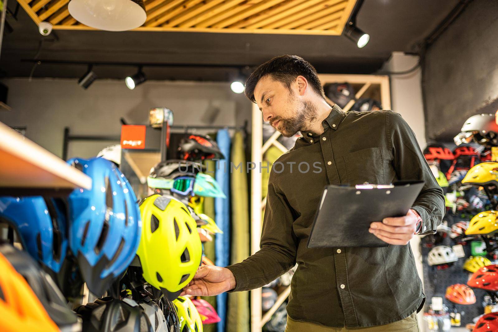 A male bike shop manager makes an inventory of sports helmets in a bike shop. The owner of a sports store with a clipboard in his hands checks the prices of bicycle helmets in the showcase.