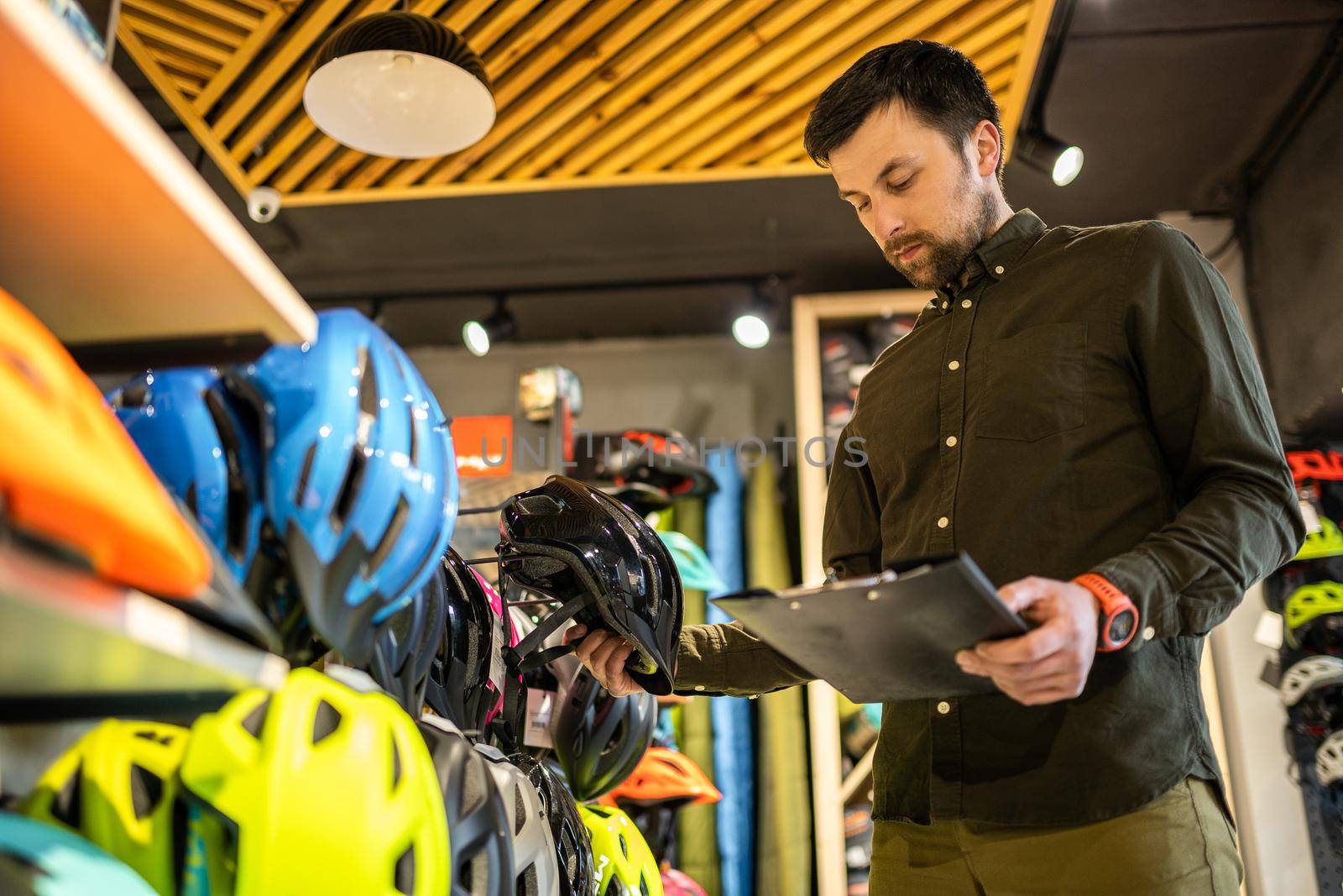 A male bike shop manager makes an inventory of sports helmets in a bike shop. The owner of a sports store with a clipboard in his hands checks the prices of bicycle helmets in the showcase by Tomashevska