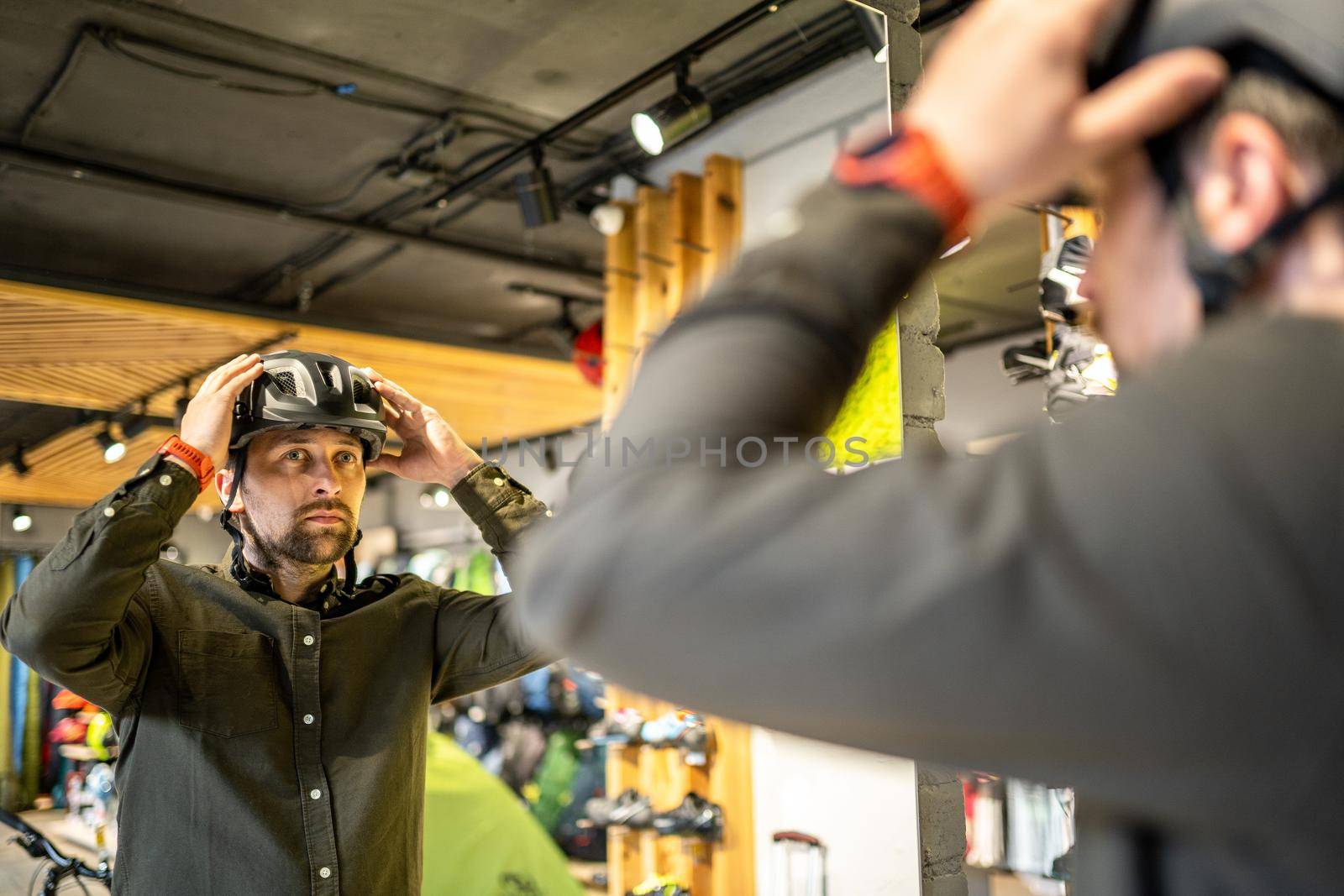 Young man came to the bicycle store. He is measuring the helmet. Male chooses helmet in sports equipment store. Purchase of new sports helmet. Customer with bicycle helmet trying on near the mirror by Tomashevska