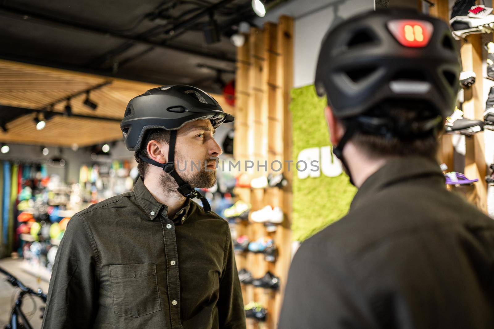 Man examining bicycle helmets in sport shop looking at herself in mirror. Trying new sports helmet in bike shop. Male putting on cycle helmet in shop. Customer in bicycle store trying on bike helmet by Tomashevska