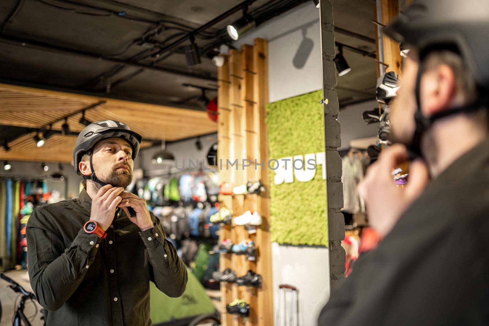 Caucasian man trying on black bicycle helmet near mirror in sporting goods store. Male buyer chooses safety helmet for cycling. Shopping in bicycle store. Person adjusts his sports helmet strap by Tomashevska
