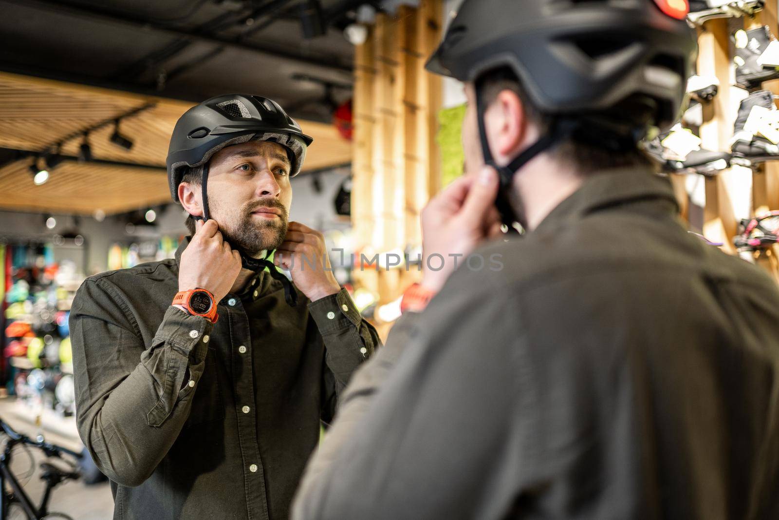 Caucasian man trying on black bicycle helmet near mirror in sporting goods store. Male buyer chooses safety helmet for cycling. Shopping in bicycle store. Person adjusts his sports helmet strap.