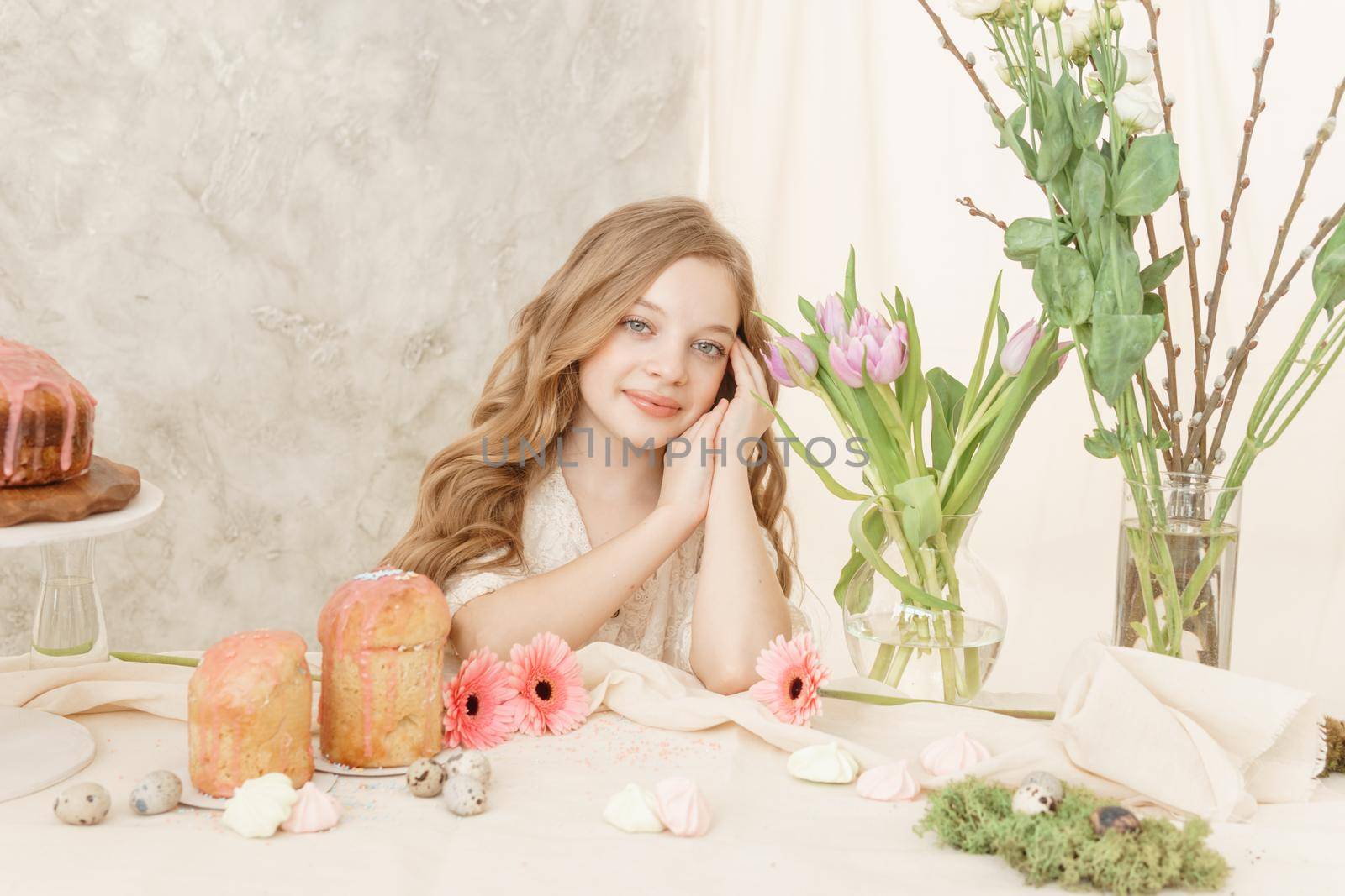 A girl with long hair in a light dress is sitting at the Easter table with cakes, spring flowers and quail eggs. Happy Easter celebration