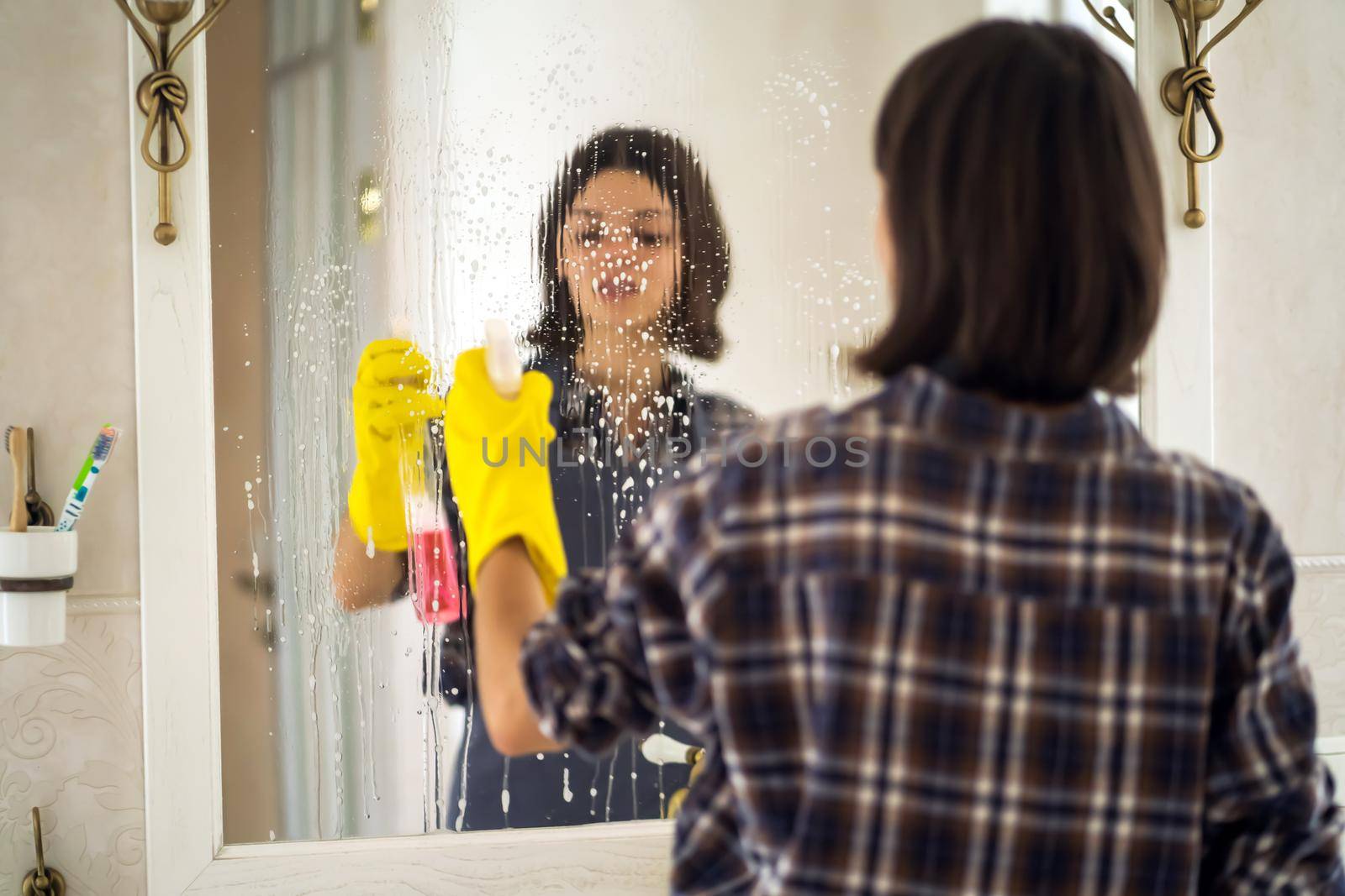 A young girl is cleaning the bathroom, applying detergent with a spray and washing the mirror with a sponge in yellow gloves on her hands. Smiling woman taking care of the cleanliness of her home.