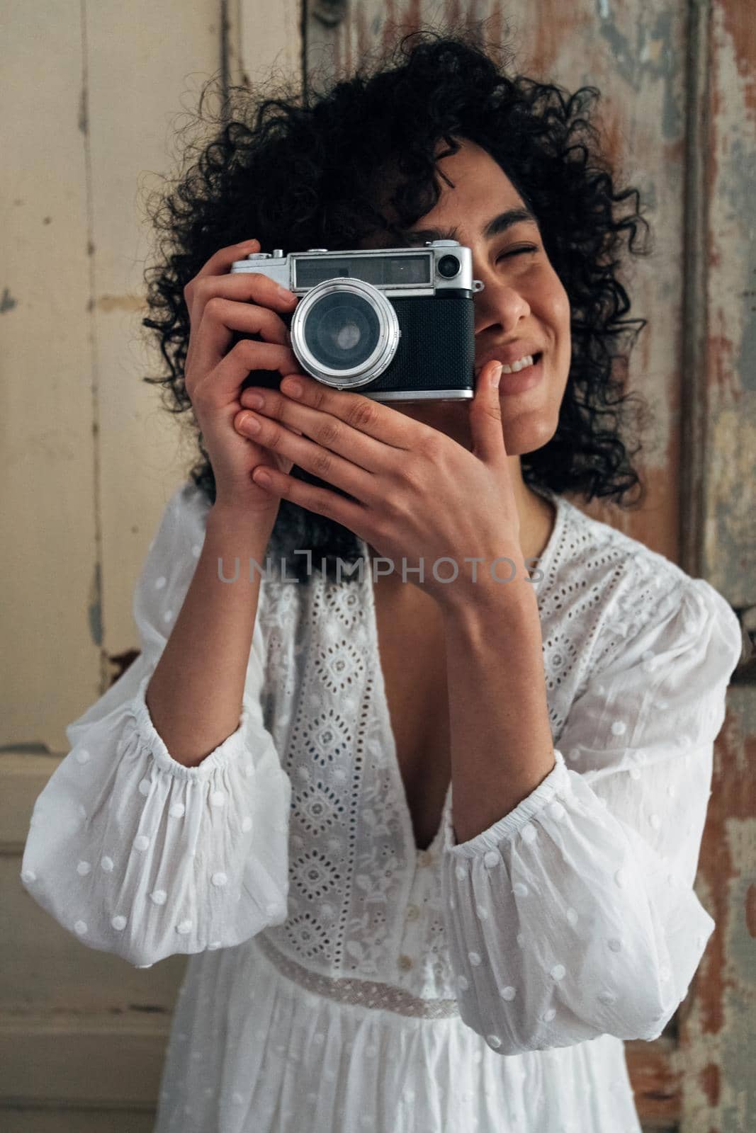 Portrait of young multiracial woman holding vintage style camera taking a picture looking at camera. Vertical image. Tourism and lifestyle concept.
