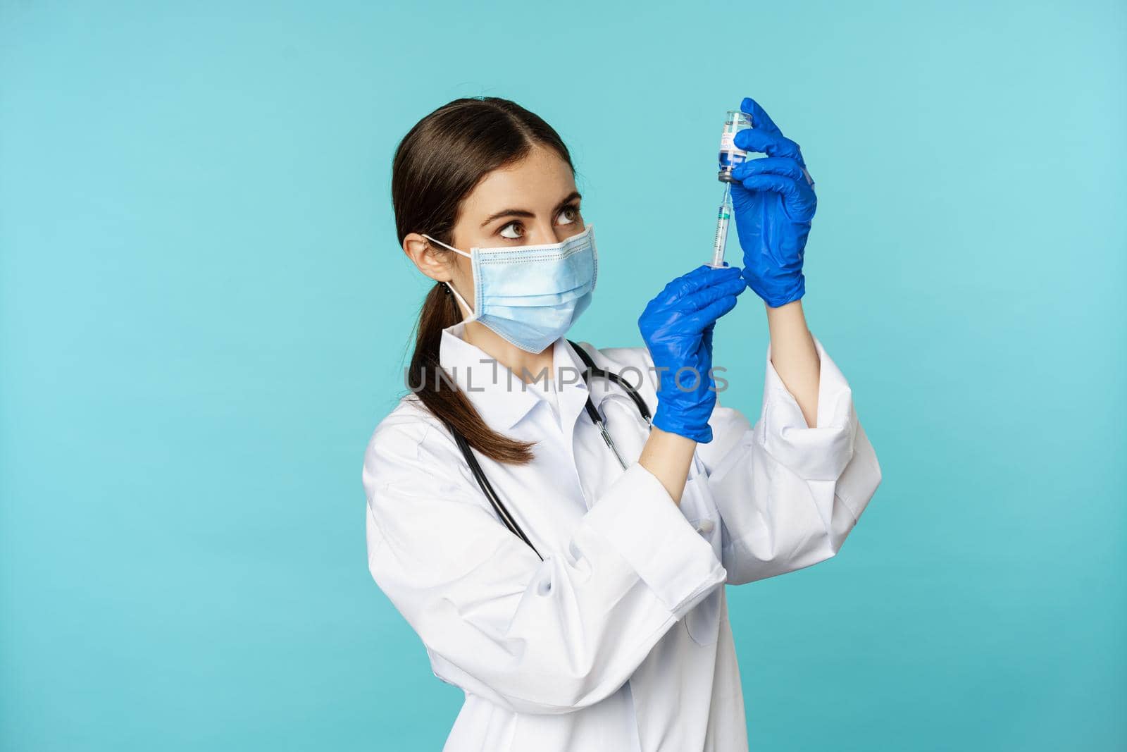 Vaccination from covid and healthcare concept. Young woman doctor, nurse in face mask and gloves, using syringe to do vaccine shot, standing over torquoise background.