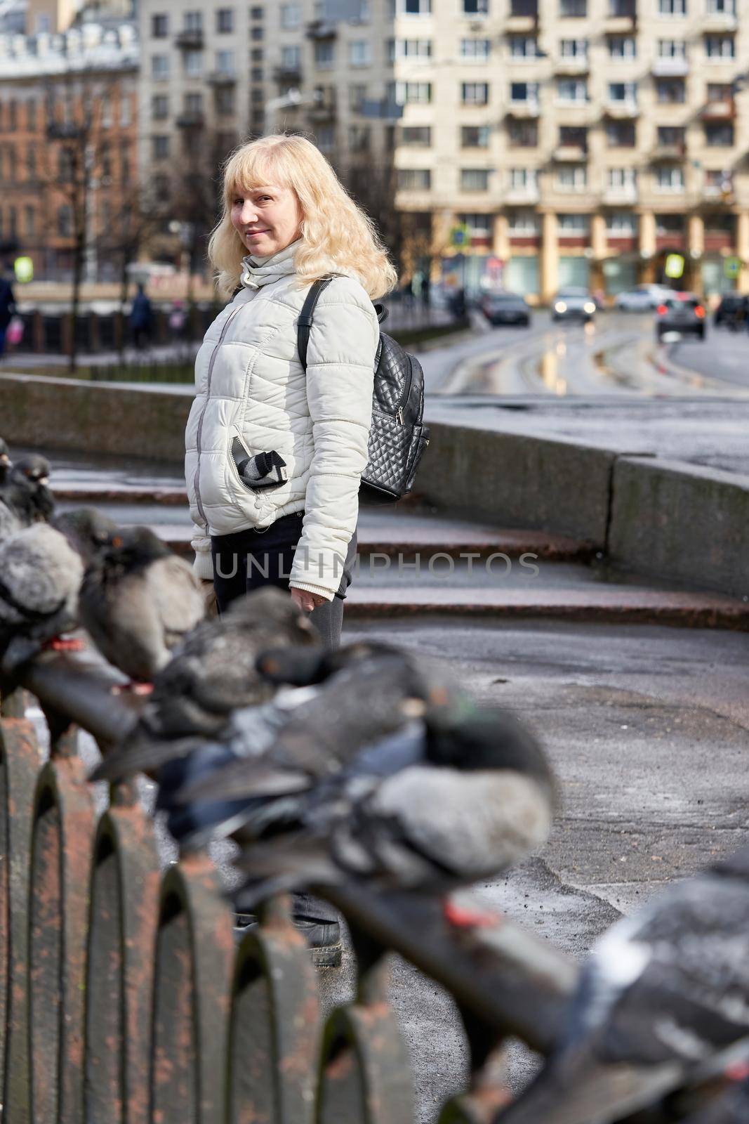 Blonde girl in a light jacket next to a metal fence and pigeons sitting on it, cityscape