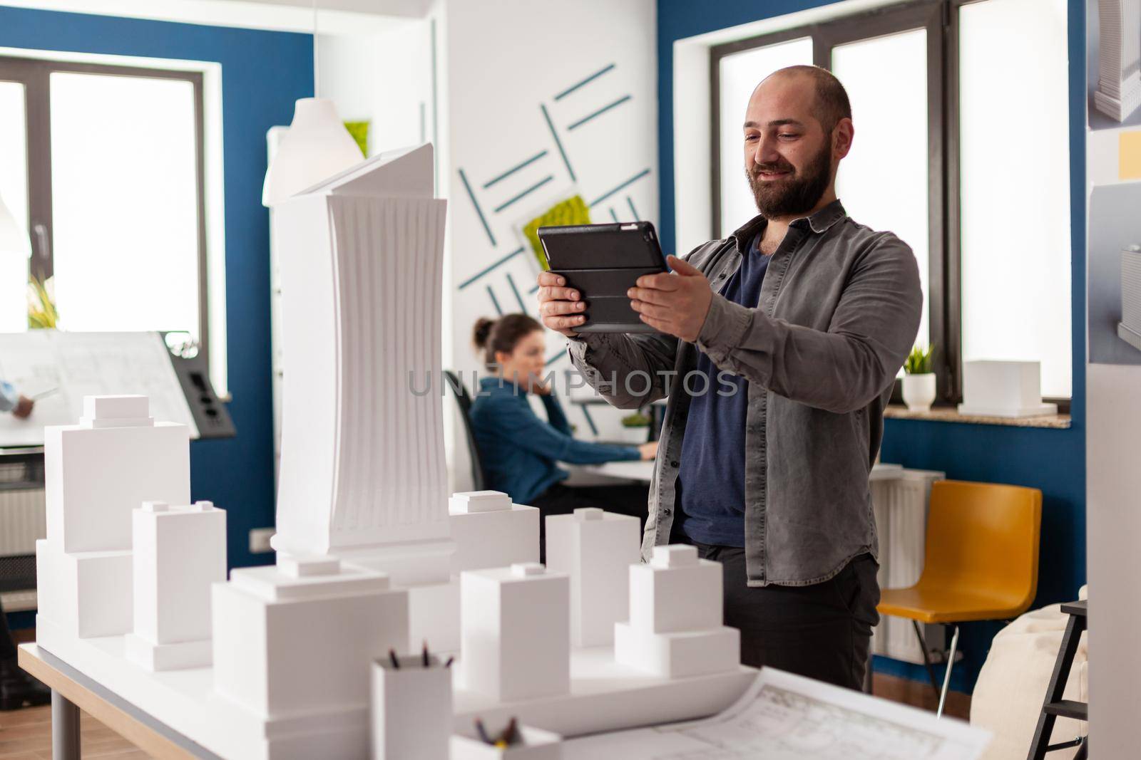 Bearded professional architect looking at tablet in front of white foam building model in architectural office. Engineer comparing blueprints on digital device with scale design of skyscraper.