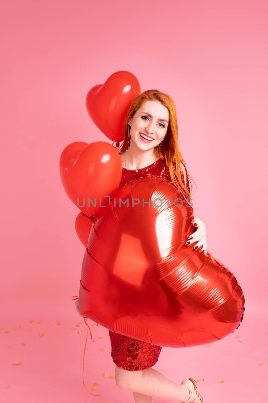 Beautiful redhead girl with red heart baloon posing. Happy Valentine's Day concept. Studio photo of beautiful ginger girl dancing on pink background.