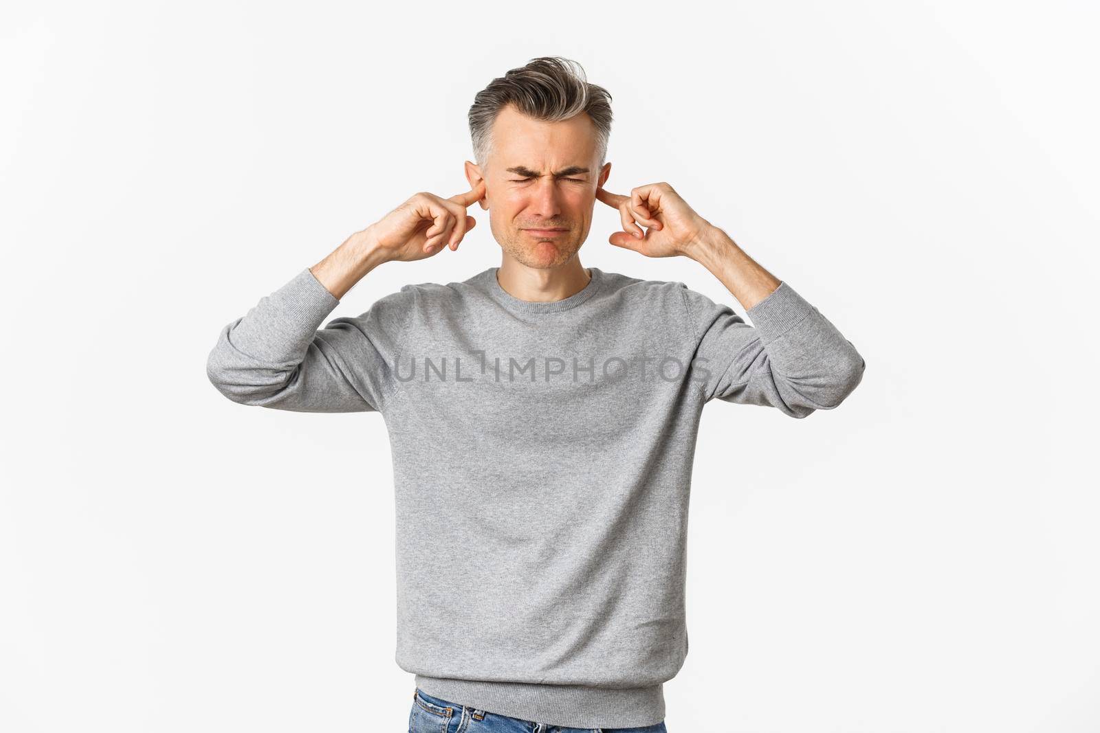 Portrait of annoyed and distressed middle-aged man, grimacing from loud disturbing noise, shut ears with fingers, standing over white background.