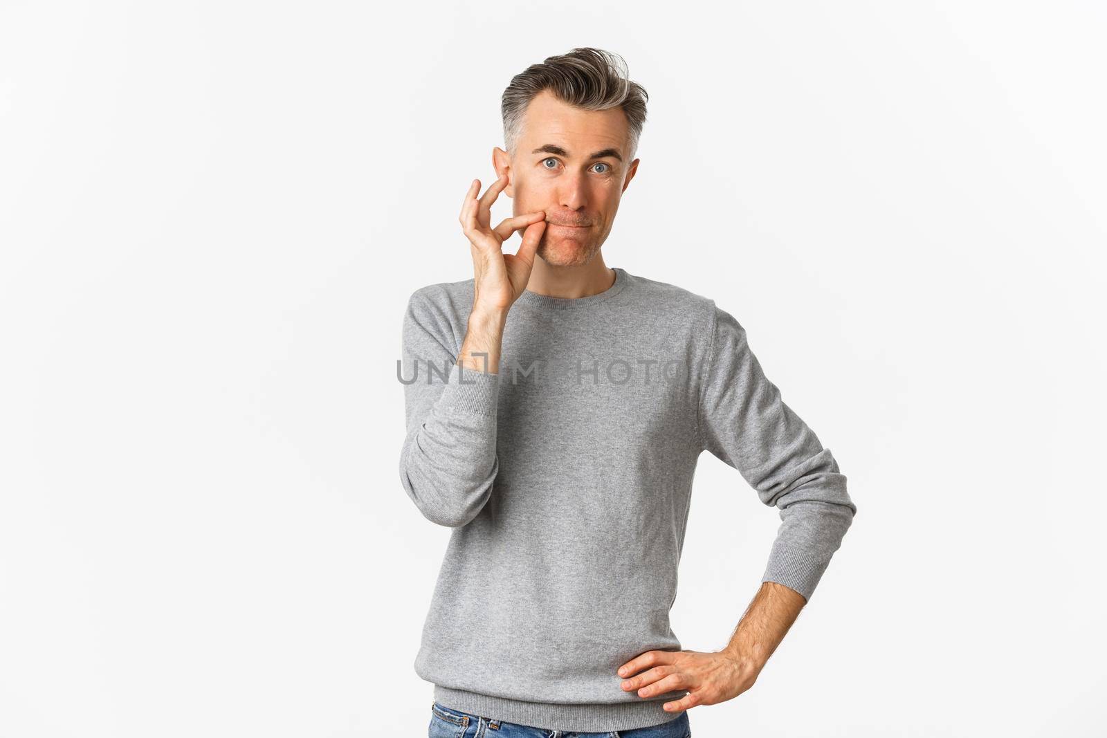 Portrait of middle-aged man making promise to keep mouth shut, zipping lips, hiding secret, standing over white background.