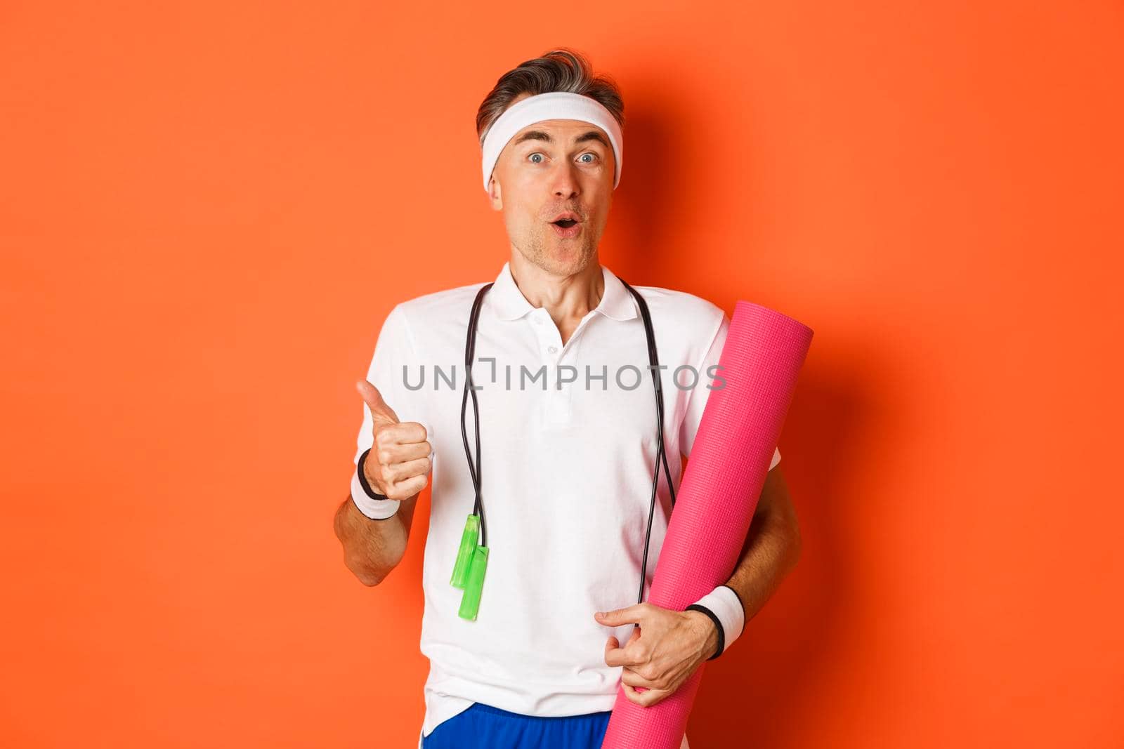 Concept of workout, gym and lifestyle. Close-up of happy and satisfied middle-aged athlete, showing thumb-up, like training session, holding yoga mat and skipping rope for fitness.