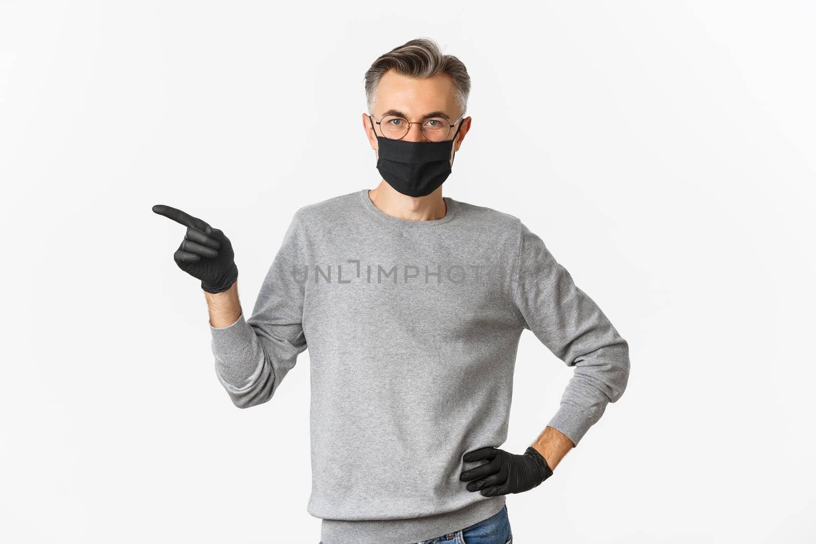 Concept of coronavirus, lifestyle and quarantine. Portrait of serious middle-aged guy in medical mask and gloves, pointing finger left at copy space, standing over white background.