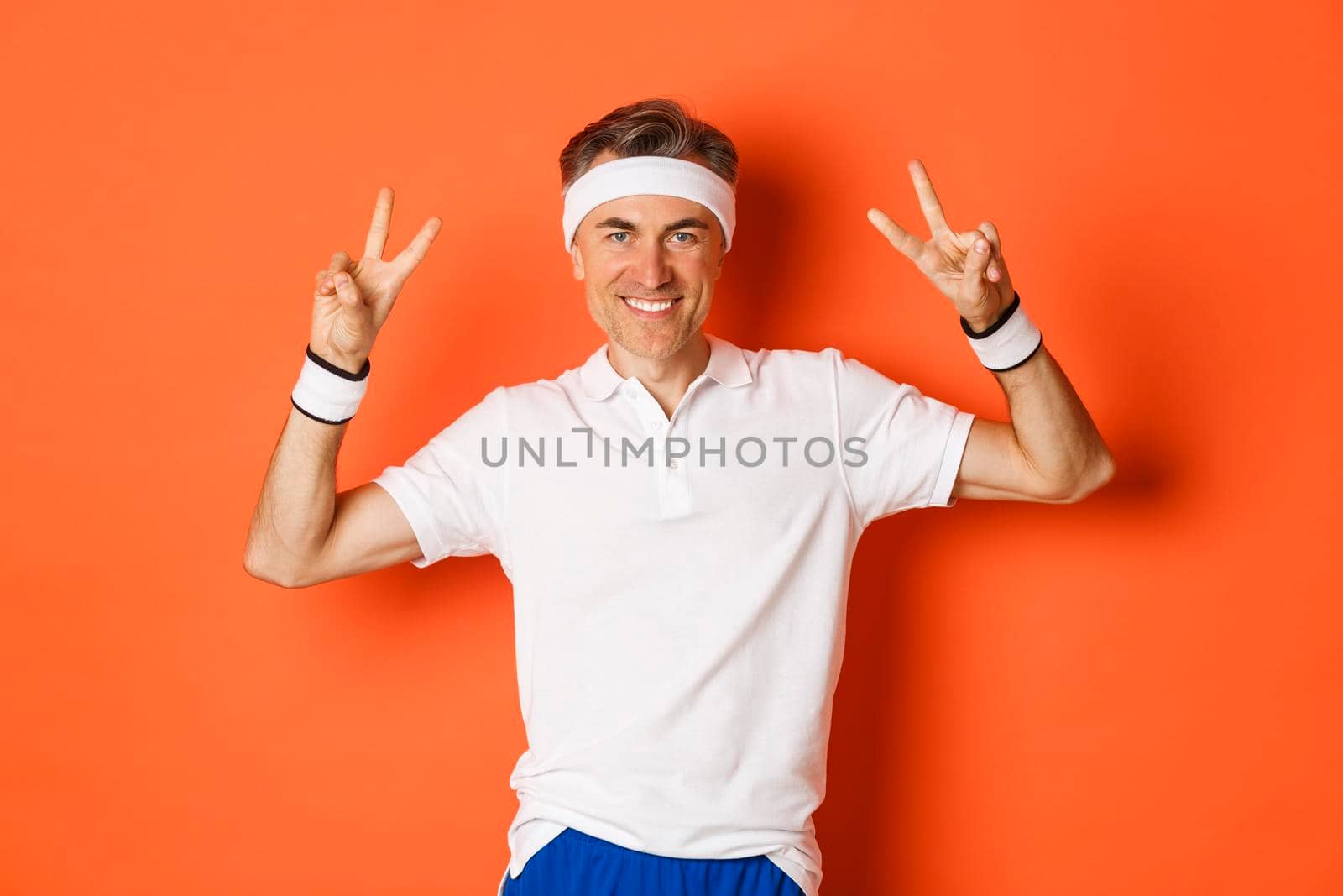 Portrait of handsome, healthy middle-aged male athlete in sportswear, showing peace signs and smiling, workout in gym, standing over orange background.