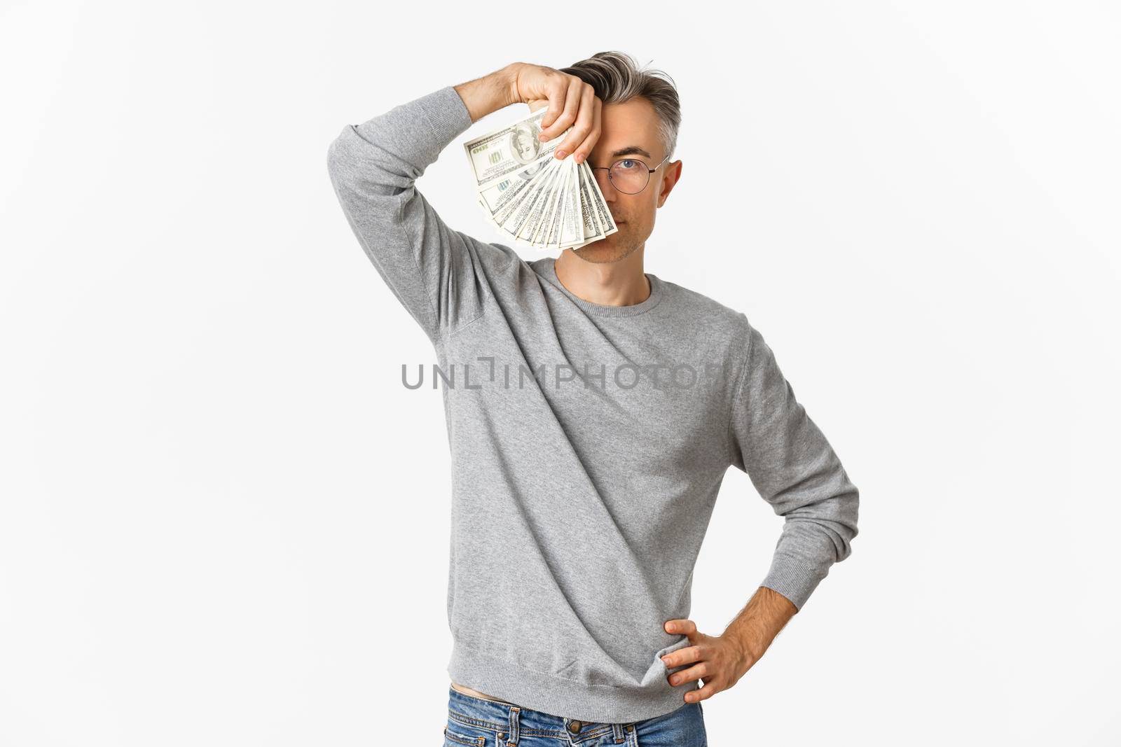 Portrait of successful and rich middle-aged man in gray sweater, showing money, standing over white background.