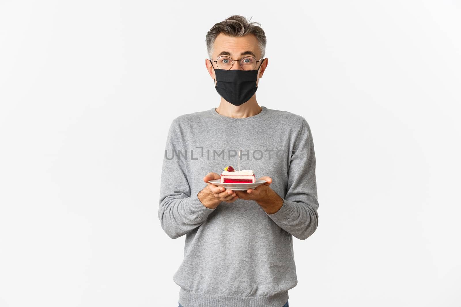 Concept of covid-19, quarantine and holidays. Portrait of amazed middle-aged man in medical mask, celebrating his birthday during coronavirus, holding b-day cake with candle.