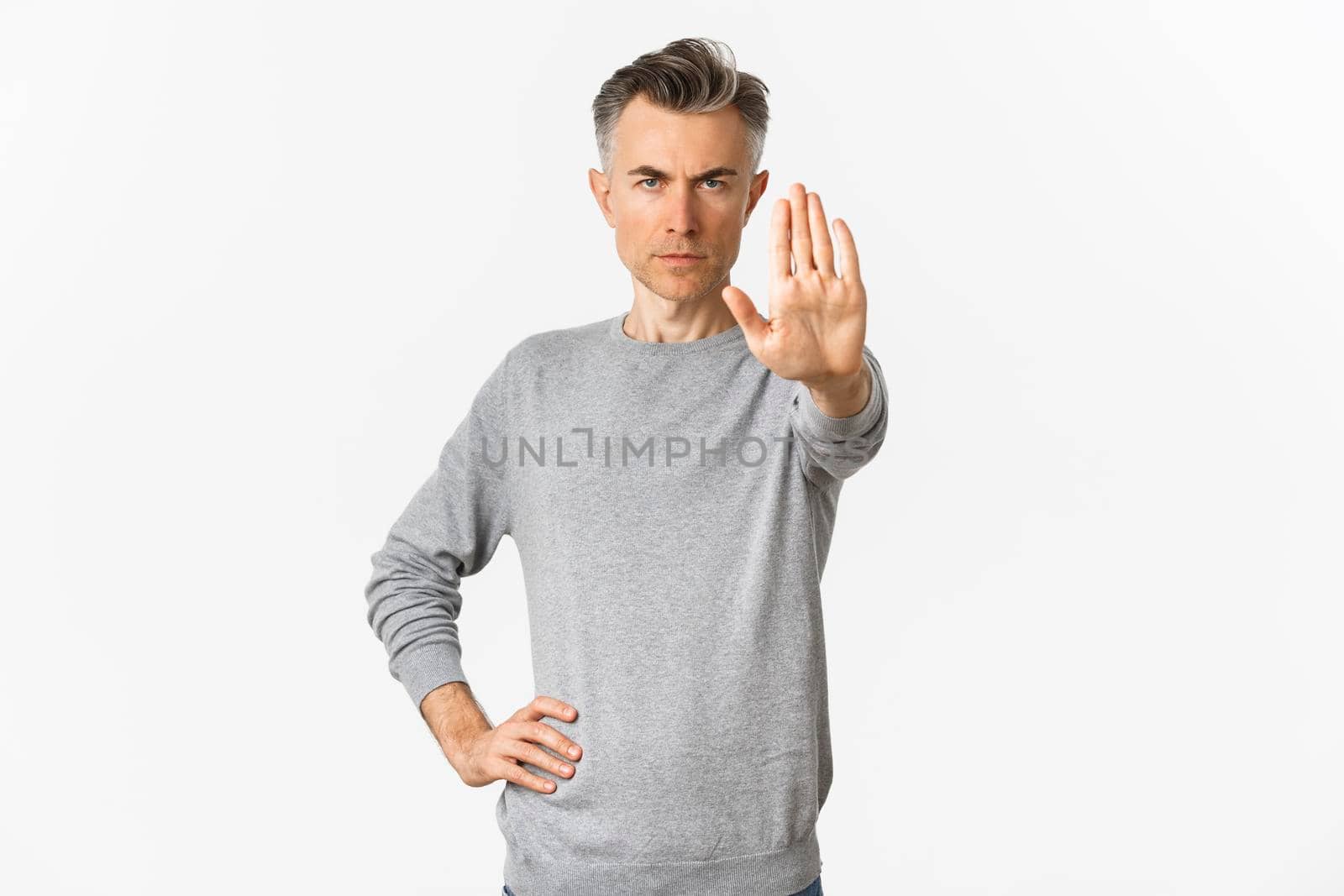 Image of serious middle-aged man extend one hand to stop you, prohibit something bad, frowning and looking with disapproval, standing over white background.